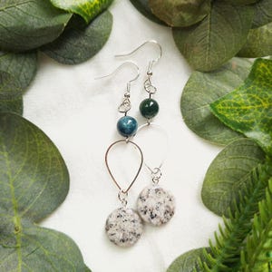 May include: A pair of dangle earrings with silver-tone hooks. Each earring features a dark green bead, a blue bead, a silver teardrop shape, and a speckled, round, light gray stone. The earrings are displayed on a white surface with green foliage.