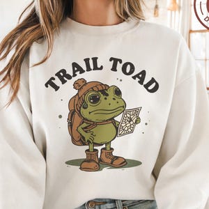 Könnte beinhalten: Cremefarbenes Sweatshirt mit einer Cartoon-Illustration einer grünen Frosch mit Rucksack, Hut und Stiefeln, der eine Karte hält. Der Text "TRAIL TOAD" ist über dem Frosch gewölbt. Ein PNG-Digitaldesign-Logo befindet sich in der oberen rechten Ecke.