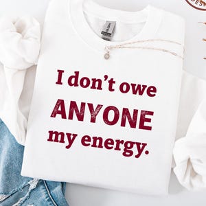 May include: A white sweatshirt with the text "I don't owe ANYONE my energy" in burgundy letters. The sweatshirt has a crew neck and long sleeves. A silver necklace with a small pendant is on the sweatshirt.