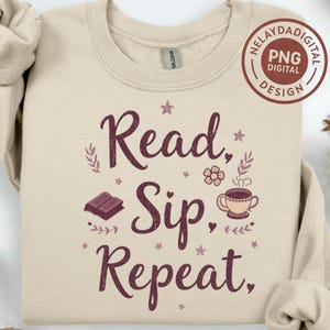 以下が含まれることがあります： クリーム色のスウェットシャツ。ダークバーガンディの筆記体で「Read, Sip, Repeat」と書かれています。デザインには本、コーヒーカップ、花のアクセントが含まれています。PNGデジタルデザインロゴが右上隅にあります。