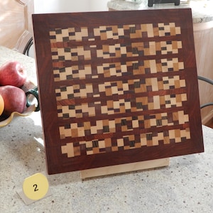 Op de afbeelding: Een vierkante houten snijplank met een donkerbruine rand en een mozaïekpatroon van lichte en donkere houten vierkanten. De plank wordt gepresenteerd op een lichtgekleurd oppervlak. Een gele sticker met het nummer 2 staat op de voorgrond.