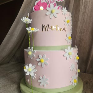 Könnte beinhalten: Zweistöckige rosa Torte, verziert mit weißen Gänseblümchen und einem grünen Sockel. Der Kuchen trägt den Namen "Martina" in goldfarbenen Buchstaben und einen Aufsatz mit der Aufschrift "You are the 1".