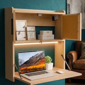 Puede incluir: Un escritorio de pared de madera clara con una superficie de trabajo plegable. El escritorio tiene un estante superior con cuadros enmarcados y cajas pequeñas. Una computadora portátil, un ratón y una pequeña planta están en el escritorio. El fondo es una pared verde azulado.