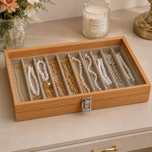 May include: A wooden jewelry box with a glass top, filled with various necklaces. The necklaces include pearl strands, gold chains, and silver chains. The box is divided into compartments with a light beige lining and a silver clasp.