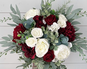 May include: A bridal bouquet featuring a mix of white, burgundy, and dark red flowers, including roses and peonies, accented with green foliage. The arrangement is held against a white wooden background.