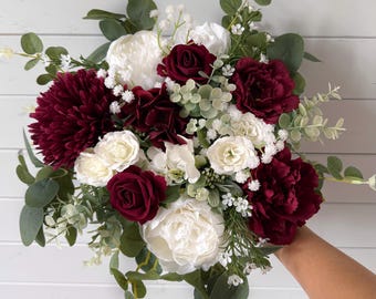 May include: A floral bouquet featuring deep burgundy and white roses, peonies, and dahlias, accented with small white baby's breath flowers and green eucalyptus leaves. The arrangement is rounded and includes various textures of faux botanical stems.