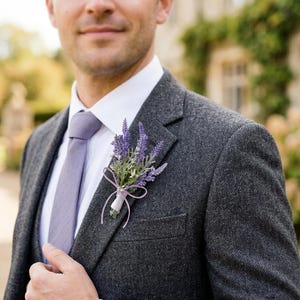 K&ouml;nnte beinhalten: Ein dunkelgrauer Tweedanzug mit wei&szlig;em Hemd und lavendelfarbener Krawatte. Eine lavendelfarbene und gr&uuml;ne Blumenansteckblume ist an der Jacke befestigt. Das Bild zeigt eine Person in formeller Kleidung.