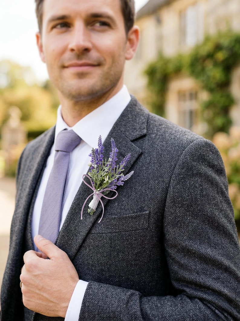 K&ouml;nnte beinhalten: Ein dunkelgrauer Tweedanzug mit wei&szlig;em Hemd und lavendelfarbener Krawatte. Eine lavendelfarbene und gr&uuml;ne Blumenansteckblume ist an der Jacke befestigt. Das Bild zeigt eine Person in formeller Kleidung.