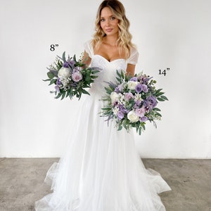 K&ouml;nnte beinhalten: Zwei Blumenstr&auml;u&szlig;e, einer mit 20 cm und der andere mit 36 cm Durchmesser, mit Lavendel-, wei&szlig;en und lila Bl&uuml;ten mit gr&uuml;nem Laub. Die Str&auml;u&szlig;e werden vor einem wei&szlig;en Kleid gehalten.