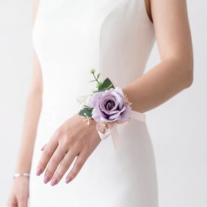 K&ouml;nnte beinhalten: Ein lila Rosen-Handgelenk-Corsage mit Gr&uuml;n und einem hellrosa Band. Das Blumengesteck wird mit einem zarten Band am Handgelenk befestigt, perfekt f&uuml;r Hochzeiten, Abschlussb&auml;lle oder besondere Anl&auml;sse.
