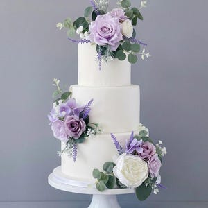 K&ouml;nnte beinhalten: Dreist&ouml;ckige wei&szlig;e Torte, verziert mit lila und wei&szlig;en Rosen, Eukalyptusbl&auml;ttern und Lavendelzweigen. Die Torte steht auf einem wei&szlig;en Sockel vor grauem Hintergrund. Eine Hochzeitstorte.
