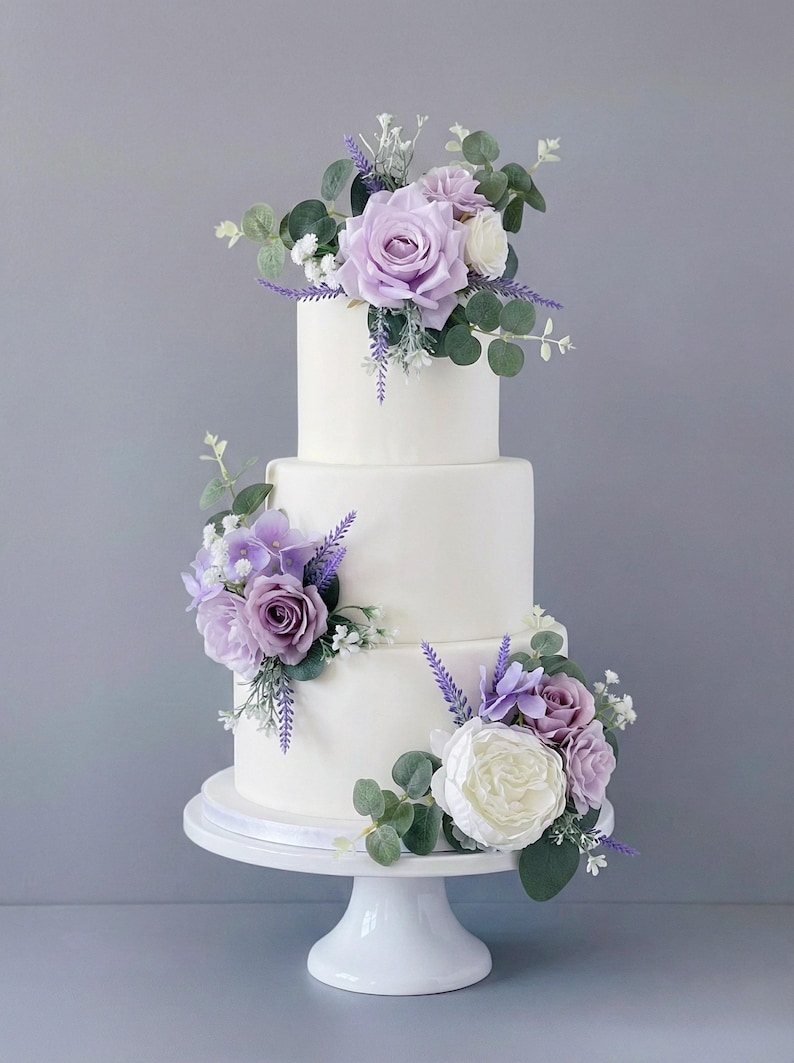 K&ouml;nnte beinhalten: Dreist&ouml;ckige wei&szlig;e Torte, verziert mit lila und wei&szlig;en Rosen, Eukalyptusbl&auml;ttern und Lavendelzweigen. Die Torte steht auf einem wei&szlig;en Sockel vor grauem Hintergrund. Eine Hochzeitstorte.