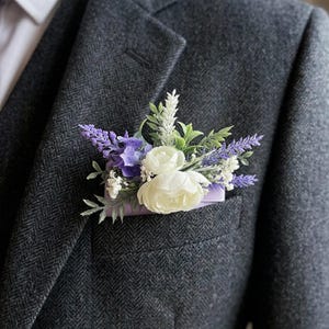 May include: A dark gray tweed suit jacket with a white dress shirt and a floral boutonniere. The boutonniere features white roses, purple lavender, and greenery, set against a lavender pocket square.