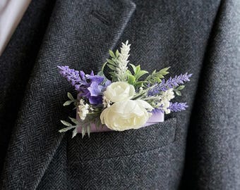 May include: A dark gray tweed suit jacket with a white dress shirt and a floral boutonniere. The boutonniere features white roses, purple lavender, and greenery, set against a lavender pocket square.