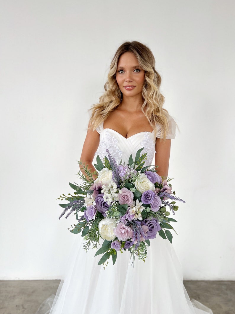 K&ouml;nnte beinhalten: Ein Brautstrau&szlig; mit einer Mischung aus Lavendel-, lila- und wei&szlig;en Blumen, darunter Rosen und Pfingstrosen, mit gr&uuml;nem Laub. Der Strau&szlig; wird vor einem wei&szlig;en Kleid gehalten.
