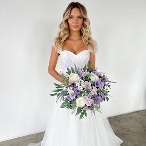 K&ouml;nnte beinhalten: Eine Braut in einem wei&szlig;en Hochzeitskleid h&auml;lt einen Strau&szlig; aus lila, Lavendel und wei&szlig;en Blumen mit gr&uuml;nem Laub. Das Kleid hat einen herzf&ouml;rmigen Ausschnitt und einen T&uuml;llrock. Der Strau&szlig; ist eine Mischung aus verschiedenen Blumenarten.