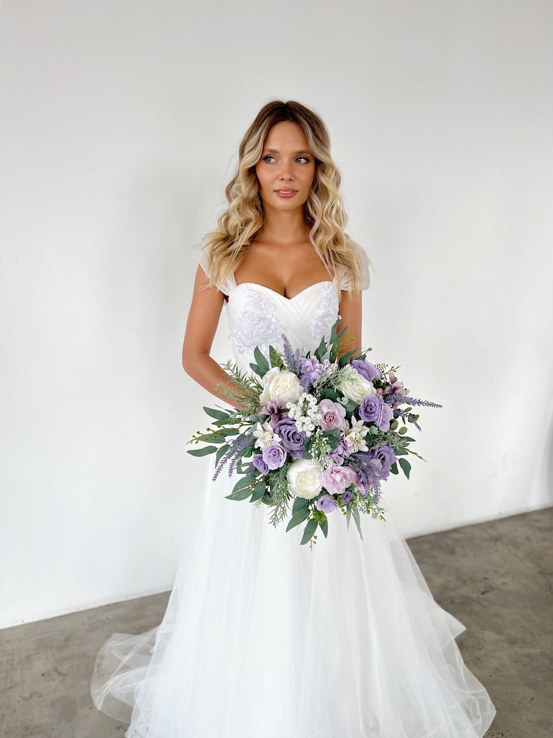 K&ouml;nnte beinhalten: Eine Braut in einem wei&szlig;en Hochzeitskleid h&auml;lt einen Strau&szlig; aus lila, Lavendel und wei&szlig;en Blumen mit gr&uuml;nem Laub. Das Kleid hat einen herzf&ouml;rmigen Ausschnitt und einen T&uuml;llrock. Der Strau&szlig; ist eine Mischung aus verschiedenen Blumenarten.
