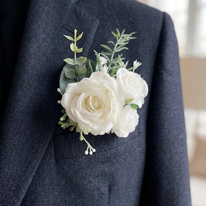 May include: A white rose boutonniere with greenery, displayed in the pocket of a dark navy blazer. The floral arrangement includes several white roses and various green leaves and sprigs, creating a classic and elegant look.