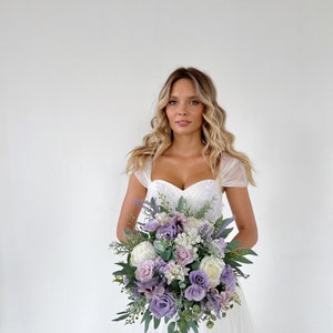 K&ouml;nnte beinhalten: Ein Brautstrau&szlig; mit Lavendel-, lila- und wei&szlig;en Rosen, akzentuiert mit Gr&uuml;n. Der Strau&szlig; wird von einer Person in einem wei&szlig;en Hochzeitskleid gehalten. Die Blumen sind rund angeordnet und erzeugen einen klassischen und eleganten Look.