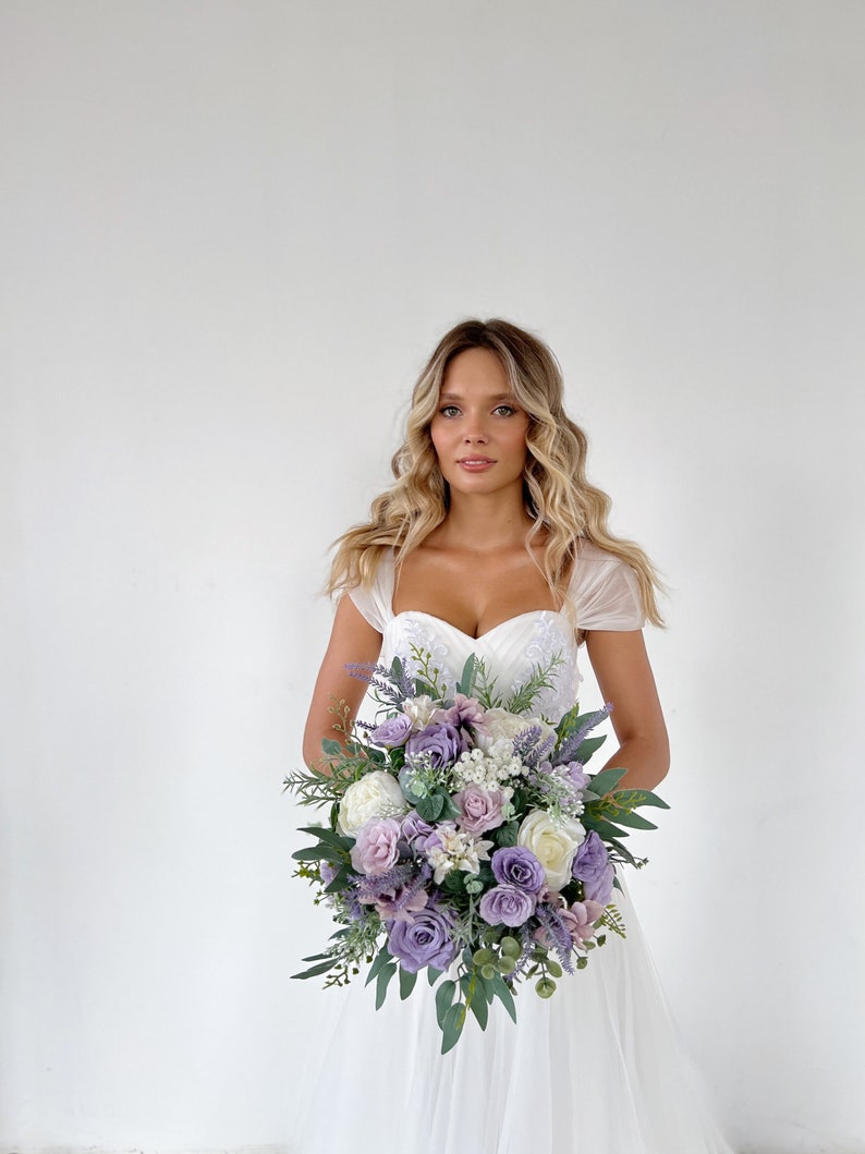 K&ouml;nnte beinhalten: Ein Brautstrau&szlig; mit Lavendel-, lila- und wei&szlig;en Rosen, akzentuiert mit Gr&uuml;n. Der Strau&szlig; wird von einer Person in einem wei&szlig;en Hochzeitskleid gehalten. Die Blumen sind rund angeordnet und erzeugen einen klassischen und eleganten Look.