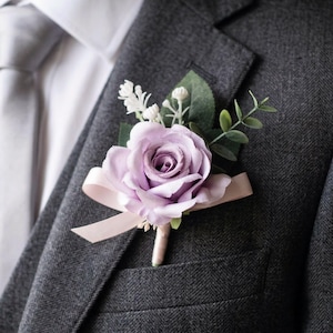 K&ouml;nnte beinhalten: Ein Lavendelrosen-Ansteckblume mit Gr&uuml;n und einem Band, an einem grauen Tweed-Anzugjacke befestigt. Die Blume ist der Mittelpunkt, mit einem hellrosa Band und Stiel. Der Anzug wird mit einem wei&szlig;en Hemd und einer grauen Krawatte kombiniert.