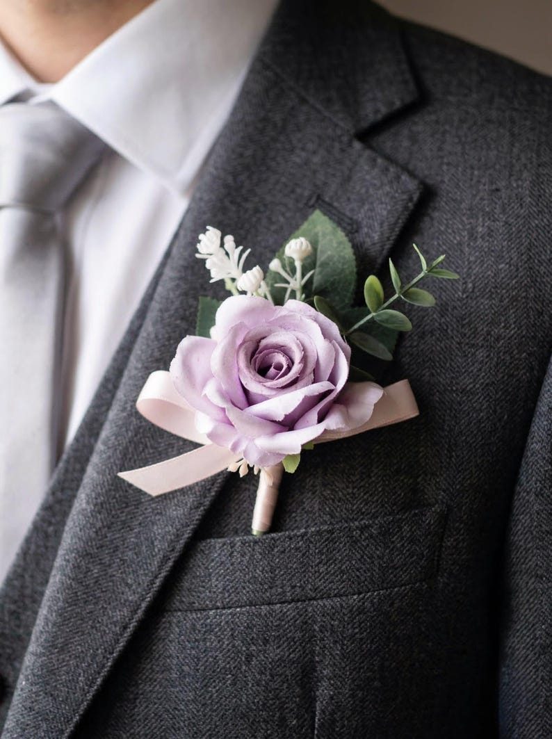 K&ouml;nnte beinhalten: Ein Lavendelrosen-Ansteckblume mit Gr&uuml;n und einem Band, an einem grauen Tweed-Anzugjacke befestigt. Die Blume ist der Mittelpunkt, mit einem hellrosa Band und Stiel. Der Anzug wird mit einem wei&szlig;en Hemd und einer grauen Krawatte kombiniert.
