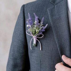 K&ouml;nnte beinhalten: Ein Ansteckblume aus Lavendel und wei&szlig;en Bl&uuml;ten mit gr&uuml;nen Bl&auml;ttern, gebunden mit einem hellvioletten Band, an einem dunkelgrauen Anzugsakko befestigt. Ein klassisches Hochzeits- oder formelles Accessoire.