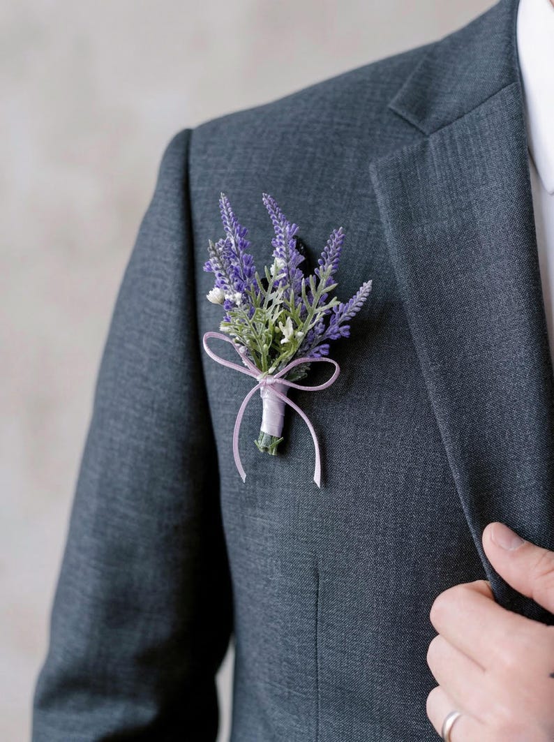 K&ouml;nnte beinhalten: Ein Ansteckblume aus Lavendel und wei&szlig;en Bl&uuml;ten mit gr&uuml;nen Bl&auml;ttern, gebunden mit einem hellvioletten Band, an einem dunkelgrauen Anzugsakko befestigt. Ein klassisches Hochzeits- oder formelles Accessoire.