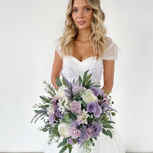 K&ouml;nnte beinhalten: Ein Brautstrau&szlig; mit Lavendel-, lila- und wei&szlig;en Blumen, darunter Rosen und Pfingstrosen, mit gr&uuml;nem Laub. Der Strau&szlig; wird vor einem wei&szlig;en Brautkleid gehalten.