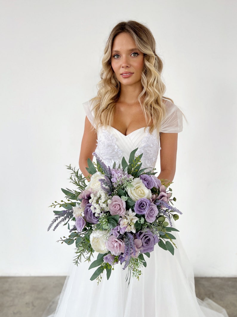 K&ouml;nnte beinhalten: Ein Brautstrau&szlig; mit Lavendel-, lila- und wei&szlig;en Blumen, darunter Rosen und Pfingstrosen, mit gr&uuml;nem Laub. Der Strau&szlig; wird vor einem wei&szlig;en Brautkleid gehalten.