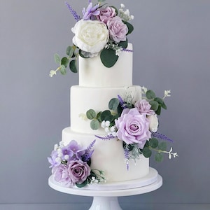 K&ouml;nnte beinhalten: Ein dreist&ouml;ckiger wei&szlig;er Kuchen, verziert mit lila und wei&szlig;en Blumen, darunter Rosen und Lavendel, und gr&uuml;nem Laub. Der Kuchen steht auf einem wei&szlig;en Tortenst&auml;nder vor grauem Hintergrund.