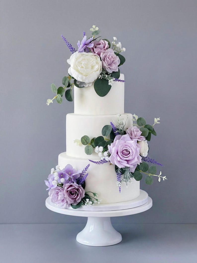 K&ouml;nnte beinhalten: Ein dreist&ouml;ckiger wei&szlig;er Kuchen, verziert mit lila und wei&szlig;en Blumen, darunter Rosen und Lavendel, und gr&uuml;nem Laub. Der Kuchen steht auf einem wei&szlig;en Tortenst&auml;nder vor grauem Hintergrund.