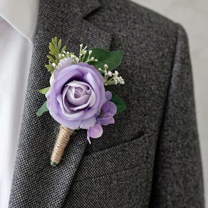 K&ouml;nnte beinhalten: Ein lila Rosen-Ansteckblume mit gr&uuml;nen Bl&auml;ttern und kleinen wei&szlig;en Bl&uuml;ten. Der Stiel ist mit Bindfaden umwickelt. Die Ansteckblume ist an einer grauen Tweedjacke befestigt.