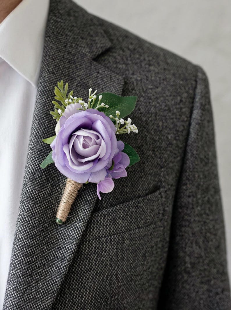 K&ouml;nnte beinhalten: Ein lila Rosen-Ansteckblume mit gr&uuml;nen Bl&auml;ttern und kleinen wei&szlig;en Bl&uuml;ten. Der Stiel ist mit Bindfaden umwickelt. Die Ansteckblume ist an einer grauen Tweedjacke befestigt.