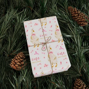 May include: A wrapped gift with white paper featuring a pattern of watercolor geese wearing pink bows, pink bows, and the text "one silly goose". The gift is tied with twine and rests on a bed of pine needles and pine cones.