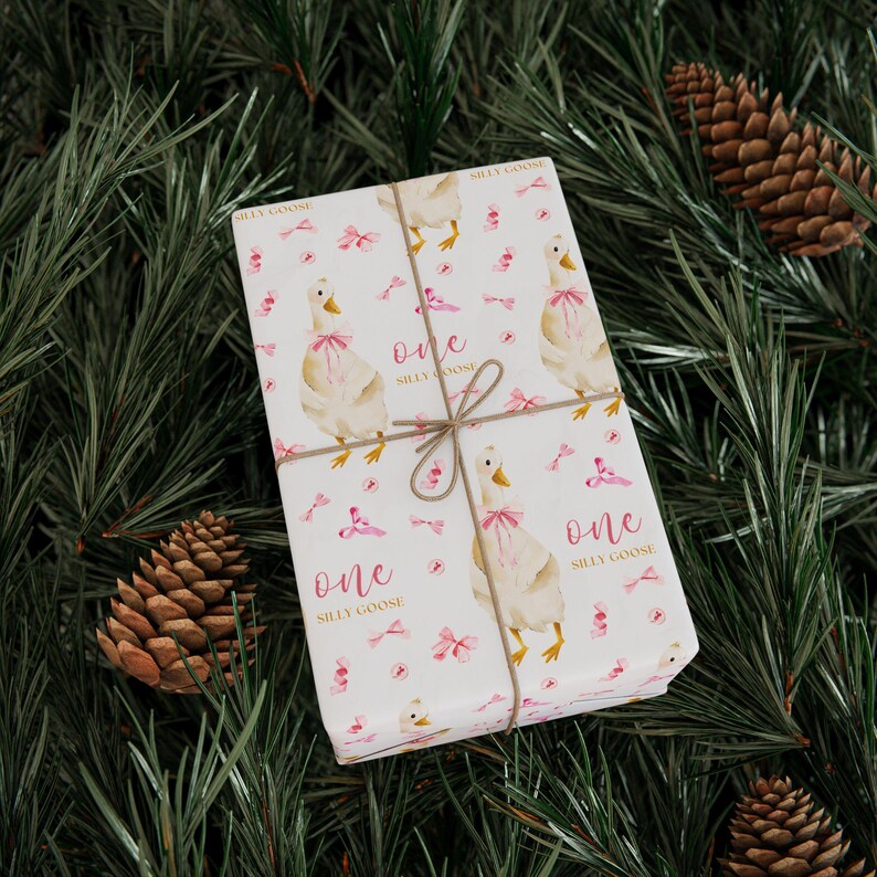 May include: A wrapped gift with white paper featuring a pattern of watercolor geese wearing pink bows, pink bows, and the text "one silly goose". The gift is tied with twine and rests on a bed of pine needles and pine cones.
