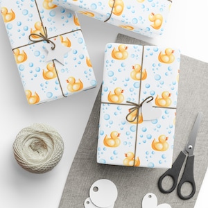 May include: Gift-wrapped presents with a white background featuring yellow rubber ducks and blue bubbles. The gifts are tied with twine and adorned with white heart-shaped tags. A ball of twine, scissors, and additional tags are also visible.