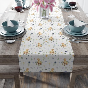 May include: A white table runner featuring a repeating pattern of yellow ducklings, blue and yellow wildflowers, and green foliage. The runner is set on a wooden table with place settings, wine glasses, and a vase of pink flowers.