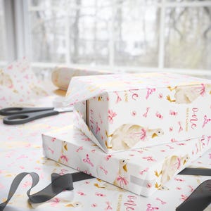 May include: Two gift boxes wrapped in white paper with a pattern of pink bows, geese, and the words "Silly Goose" and "one." A pair of scissors and a roll of wrapping paper are visible in the background. A black ribbon is in the foreground.