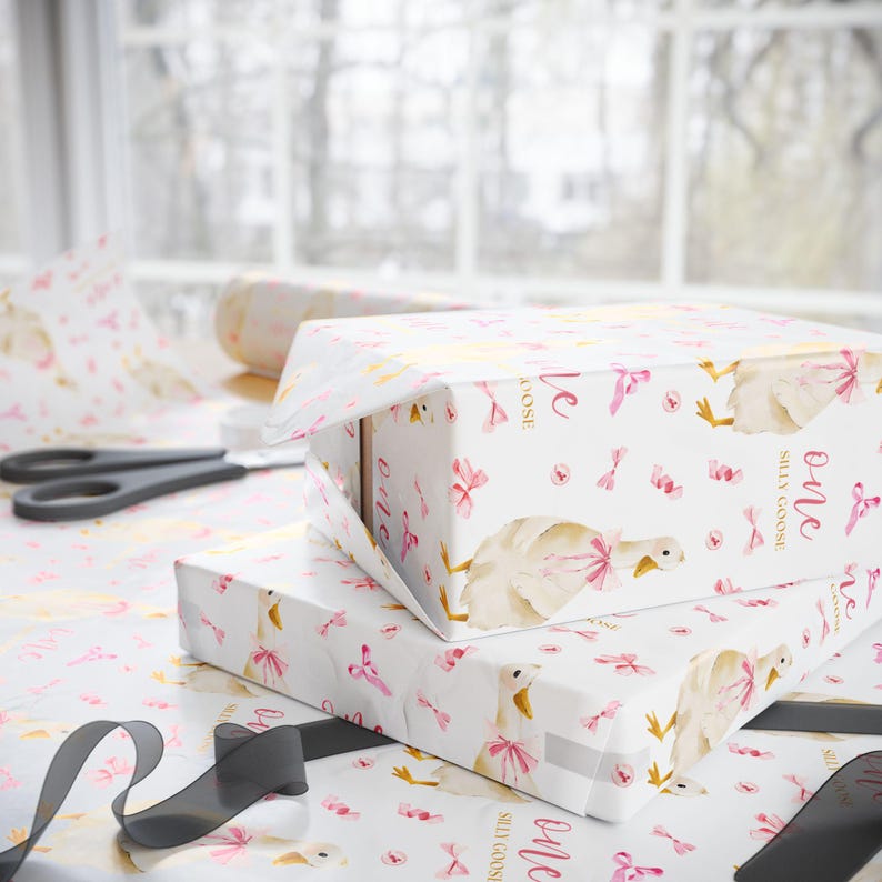 May include: Two gift boxes wrapped in white paper with a pattern of pink bows, geese, and the words "Silly Goose" and "one." A pair of scissors and a roll of wrapping paper are visible in the background. A black ribbon is in the foreground.