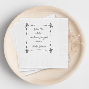 May include: White square napkins with the text "For this child we have prayed" and "Baby Johnson July 2026" in black script. The napkins are stacked on a light wooden plate with a sprig of dried flowers.