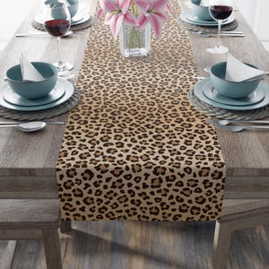 Può includere: Una tovaglia con stampa leopardata nei toni del marrone e del beige. La tovaglia è posizionata su un tavolo di legno, completata da stoviglie, calici e un vaso di fiori rosa. La tovaglia aggiunge un tocco selvaggio alla sala da pranzo.