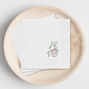 May include: White square napkins with a decorative border, stacked on a wooden plate. Each napkin features a green and brown illustration of a smiling potted plant with the text "Baby Johnson".