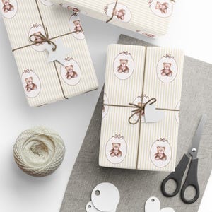 May include: Wrapped gifts with a cream and beige striped pattern featuring teddy bear illustrations in oval frames. Each package is tied with twine and a white tag. A ball of twine, scissors, and blank tags are also present.