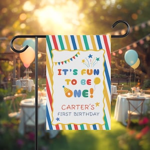 May include: A rectangular garden flag featuring a colorful striped border in red, yellow, blue, and green. The center displays the text "IT'S FUN TO BE ONE!" with a party hat, balloon, and star graphics, followed by "CARTER'S FIRST BIRTHDAY".