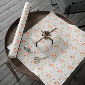 May include: White wrapping paper with a repeating pattern of cartoon ducks, balloons, and small gift boxes in peach, pink, and gold. A wrapped gift with a brown ribbon, scissors, and tape are on the paper. A roll of the same paper is also visible.