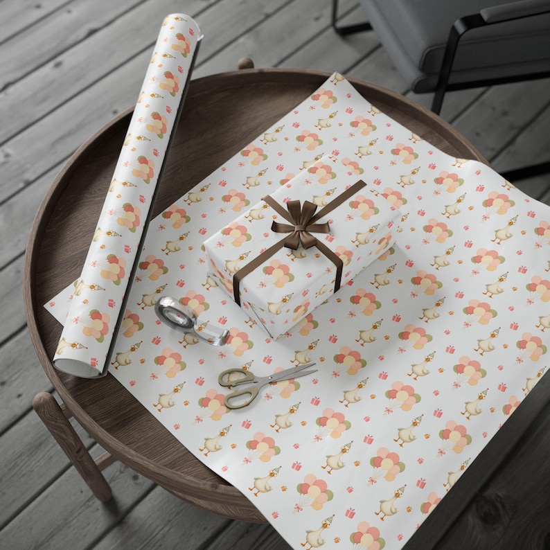 May include: White wrapping paper with a repeating pattern of cartoon ducks, balloons, and small gift boxes in peach, pink, and gold. A wrapped gift with a brown ribbon, scissors, and tape are on the paper. A roll of the same paper is also visible.