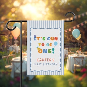 May include: A rectangular garden flag with a light blue and white vertical striped border. The center features a white background with a scalloped blue frame, the text "IT'S FUN TO BE ONE!" in colorful letters, and "CARTER'S FIRST BIRTHDAY" below.
