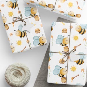 Puede incluir: Papel de regalo blanco con un patrón repetitivo de abejas de dibujos animados, tarros de miel, flores y soles. Las abejas son amarillas y negras con alas azules. Los tarros de miel son marrones con una tapa amarilla y la palabra "Honey" escrita en ellos. Las flores son amarillas y naranjas. Los soles son amarillos con rayos naranjas. El papel está atado con cuerda y una etiqueta blanca.