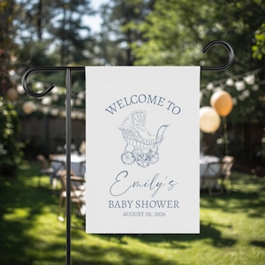 May include: A white garden flag with the text "WELCOME TO Emily's BABY SHOWER AUGUST 20, 2026" in blue. A vintage baby carriage illustration is above the text. The flag is hanging on a black metal stand in a grassy yard.